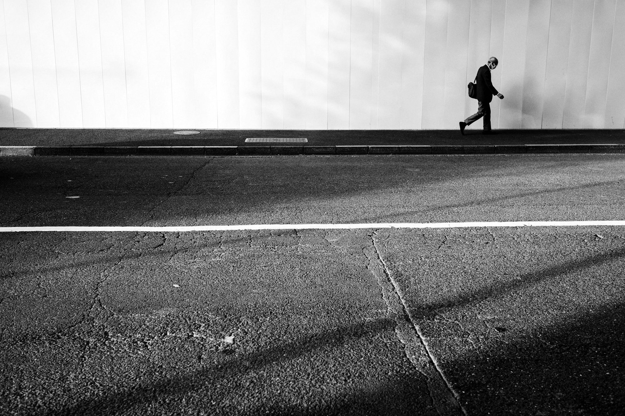Un homme en costume longe une palissade blanche ; une ligne blanche traverse la chaussée vide sous lui